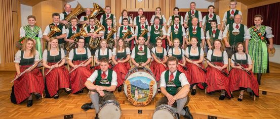 A brass band dressed in green and red poses for a photo. The band consists of various wind and percussion instruments. A drum with a town logo is in the center.