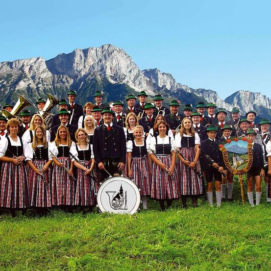 Eine Musikband in traditioneller Kleidung steht auf einer Wiese mit Bergen im Hintergrund. Die Band besteht aus männlichen und weiblichen Mitgliedern, die verschiedene Musikinstrumente halten.