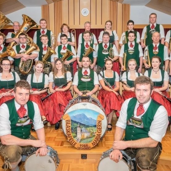 Eine große Gruppe von Musikern in Grün und Rot posiert für ein Foto. Sie halten verschiedene Instrumente wie Trompeten, Posaunen und Trommeln. Ein Schlagzeug mit der Aufschrift Blaskapelle Maria Gern steht vor ihnen. Hinter ihnen ist eine Uhr an der Wand angebracht.