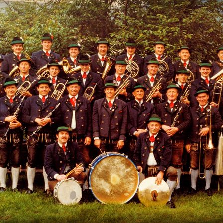 Eine Gruppe von Musikern, gekleidet in traditioneller Kleidung mit grünen Hüten, posiert für ein Foto. Sie halten verschiedene Instrumente wie Trompeten, Posaunen und Trommeln. Der Ort ist draußen, mit Bäumen im Hintergrund.