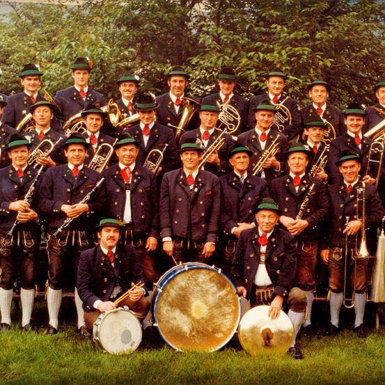 Eine Gruppe von Musikern, gekleidet in traditioneller Kleidung mit grünen Hüten, posiert für ein Foto. Sie halten verschiedene Instrumente wie Trompeten, Posaunen und Trommeln. Der Ort ist draußen, mit Bäumen im Hintergrund.