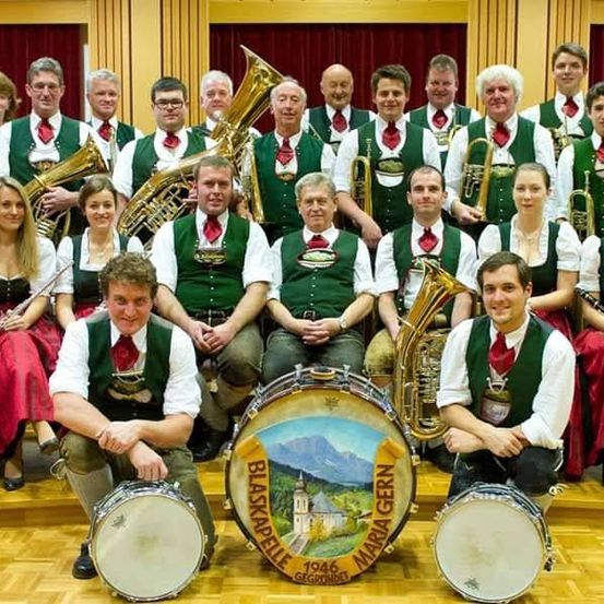 Eine Gruppe von Musikern, die grüne Westen mit roten Krawatten tragen, darunter Männer und Frauen, posieren mit ihren Instrumenten vor einem roten Vorhang für ein Foto.