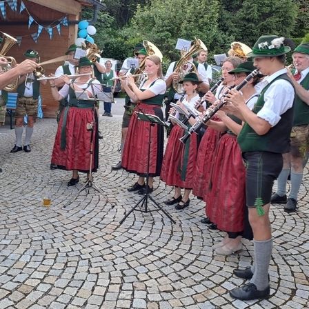 Eine Gruppe von Musikern in traditioneller bayerischer Kleidung spielt auf einem gepflasterten Platz. Sie spielen verschiedene Instrumente, darunter Trompeten, Tuben und Klarinetten.
