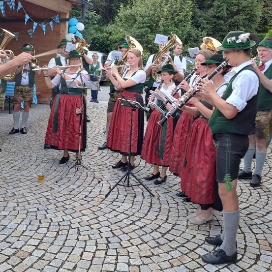 Eine Gruppe von Musikern in traditioneller bayerischer Kleidung spielt auf einem gepflasterten Platz. Sie spielen verschiedene Instrumente, darunter Trompeten, Tuben und Klarinetten.