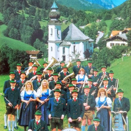 Eine Gruppe von Musikern in traditioneller bayerischer Kleidung posiert für ein Foto in einem malerischen Dorf mit Bergen und einer Kirche im Hintergrund.