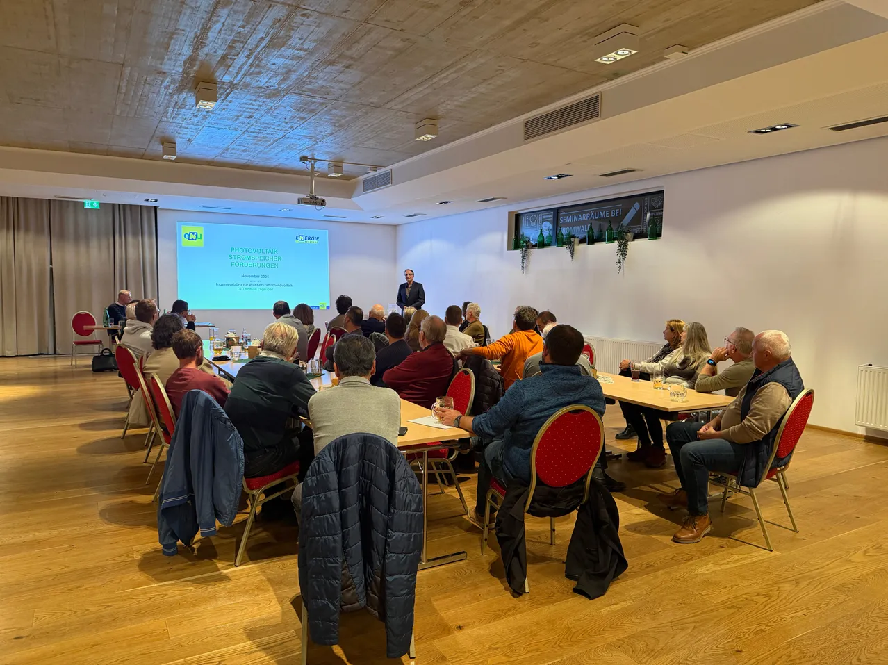 Ein Seminar mit einem Redner, der an einer Projektionsleinwand in einem Raum mit Holzböden und Tischen präsentiert, wo die Teilnehmer auf Stühlen sitzen.
