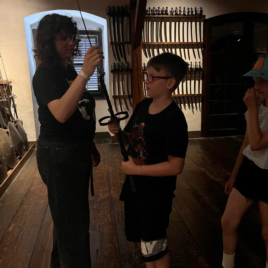 Eine Frau zeigt einem Jungen ein Schwert in einem Museum. Hinter ihnen steht ein Mädchen. Sie befinden sich in einem Raum mit Holzböden und Wänden, die mit zahlreichen Schwertern geschmückt sind.