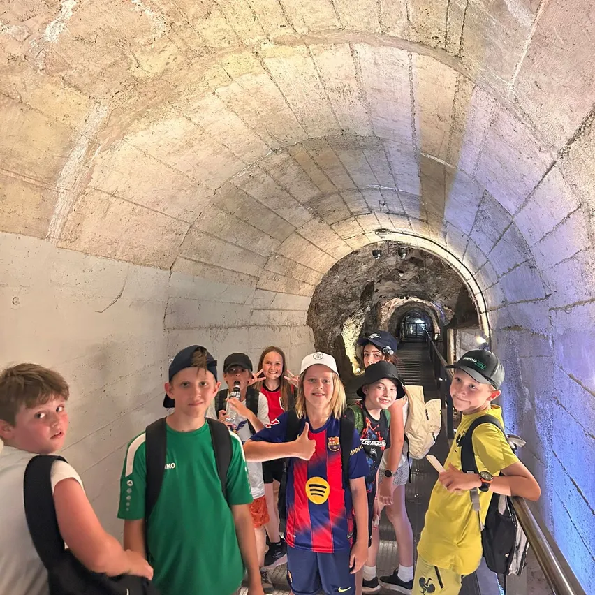 Eine Gruppe junger Leute geht durch einen gewölbten Tunnel. Sie tragen Mützen und haben Rucksäcke. Einige posieren für ein Foto. Der Tunnel ist aus Stein.