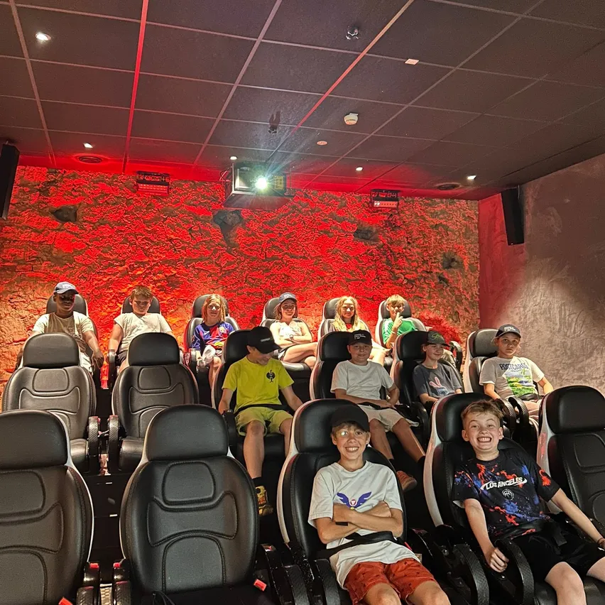 Mehrere Kinder in einem Kino, die in Sesseln sitzen, Brillen, Mützen und Turnschuhe tragen, einige lächeln. Eine rote Wand hinter ihnen und ein Projektor an der Decke.