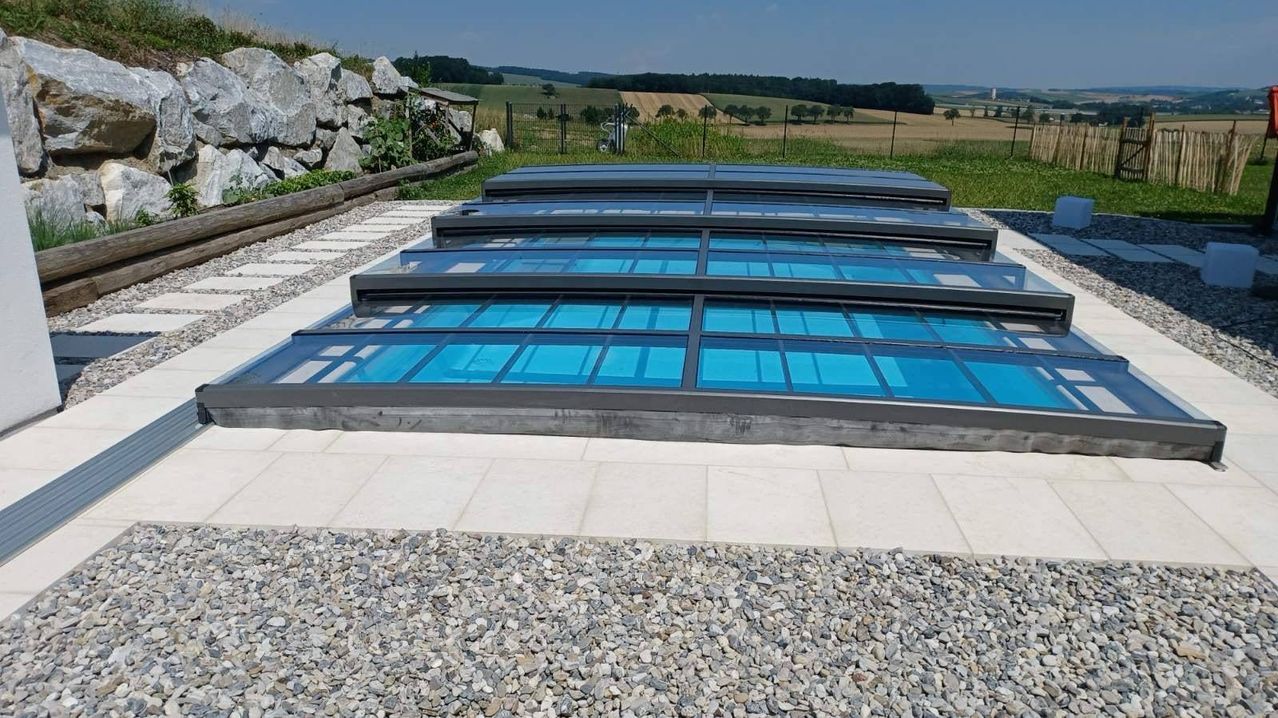 Ein Glasdachpool steht im Freien. Er hat klare Glasscheiben. Um ihn herum liegt Kies. Dahinter befindet sich eine Rasenfläche mit einer Steinmauer.