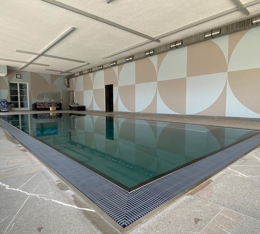Ein Hallenbad mit einer Glasbarriere und einem gefliesten Boden. Die Wände haben ein geometrisches Muster, und in der Ecke steht eine Couch.