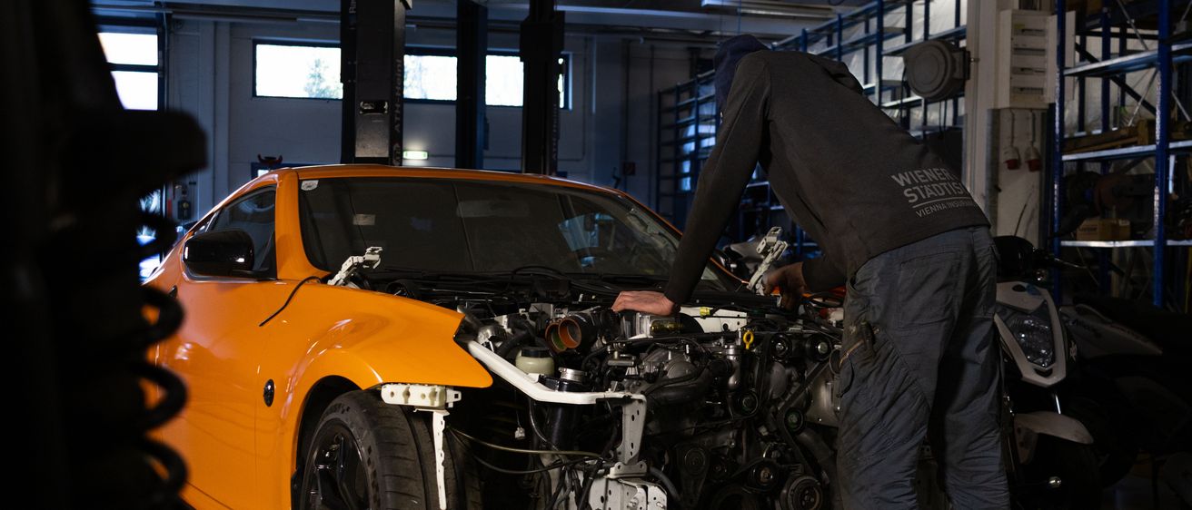 Eine Person arbeitet an einem orangefarbenen Auto in einer Garage. Der Wagen steht auf einer Plattform.
