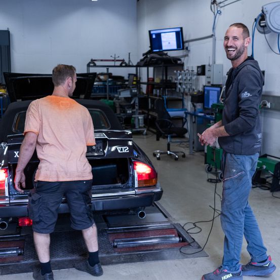 Zwei Männer in einer Werkstatt, einer lächelt, der andere arbeitet an einem Auto. Im Hintergrund sind verschiedene Werkzeuge und Geräte zu sehen.