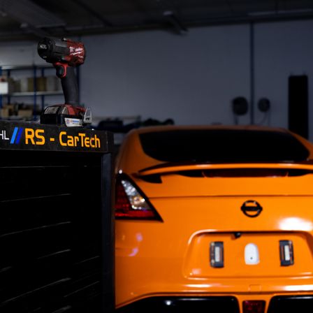 Ein orangefarbenes Auto steht in einer Garage, mit einem Werkzeugkasten und einer Bohrmaschine auf dem Schriftzug 'RS - CarTech'. Die Rücklichter des Autos sind an.