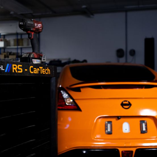 Ein orangefarbenes Auto steht in einer Garage, mit einem Werkzeugkasten und einer Bohrmaschine auf dem Schriftzug 'RS - CarTech'. Die Rücklichter des Autos sind an.