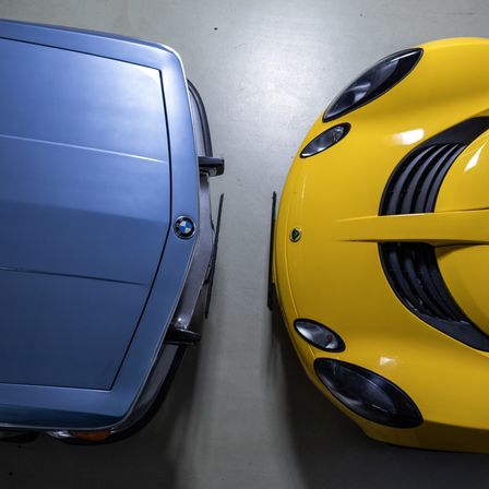 Ein Nahaufnahme der Rückseite eines blauen BMW und der Front eines gelben Sportwagens mit schwarzen Akzenten. Beide Autos sind auf einem weißen Boden geparkt.