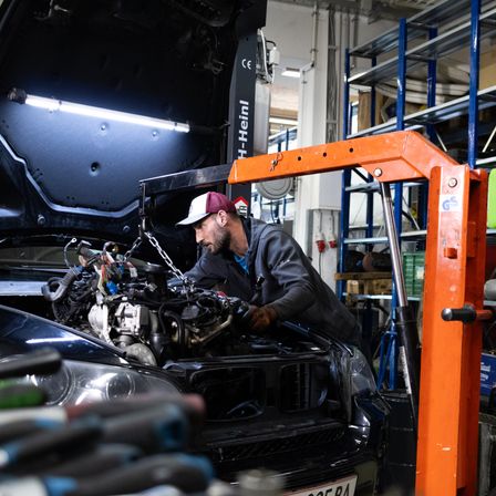 Ein Mechaniker arbeitet in einer Werkstatt an einem Auto. Er trägt eine Kappe und Handschuhe und hält ein Werkzeug. Die Motorhaube des Autos ist geöffnet und der Motor ist sichtbar.