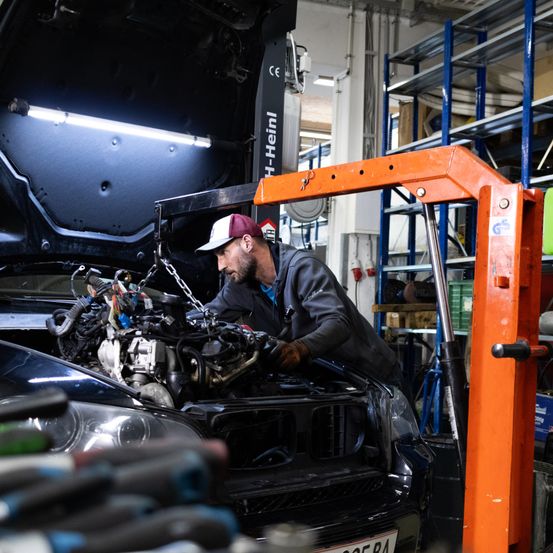 Ein Mechaniker arbeitet in einer Werkstatt an einem Auto. Er trägt eine Kappe und Handschuhe und hält ein Werkzeug. Die Motorhaube des Autos ist geöffnet und der Motor ist sichtbar.