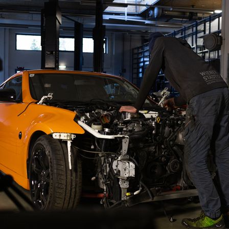 Ein Mechaniker arbeitet an einem orangefarbenen Sportwagen mit offenem Motorhauben in einer Garage. Der Wagen hat seine vorderen Reifen angehoben und der Motor ist freigelegt.