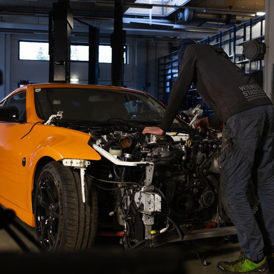 Ein Mechaniker arbeitet an einem orangefarbenen Sportwagen mit offenem Motorhauben in einer Garage. Der Wagen hat seine vorderen Reifen angehoben und der Motor ist freigelegt.