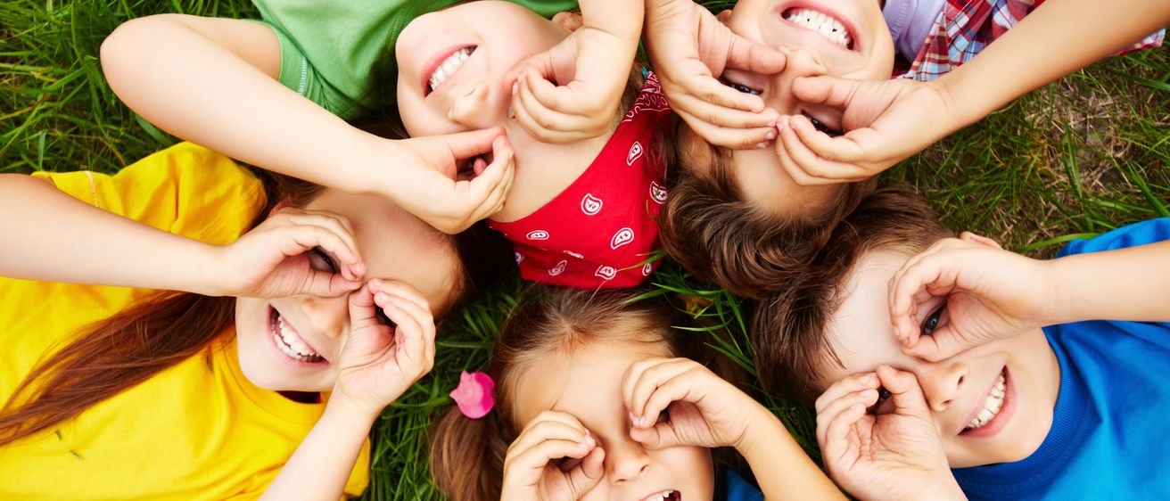 Eine Gruppe junger Kinder liegt auf dem Gras, lächelt und posiert für die Kamera. Sie haben ihre Finger auf den Augen und bilden Herzformen.