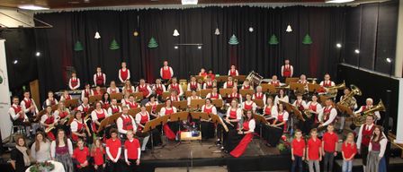 Ein großes Orchester mit verschiedenen Instrumenten tritt auf einer Bühne auf. Die Bandmitglieder sind in roten und weißen Kostümen gekleidet. Die Bühne ist mit Weihnachtsbäumen und Lichtern geschmückt.