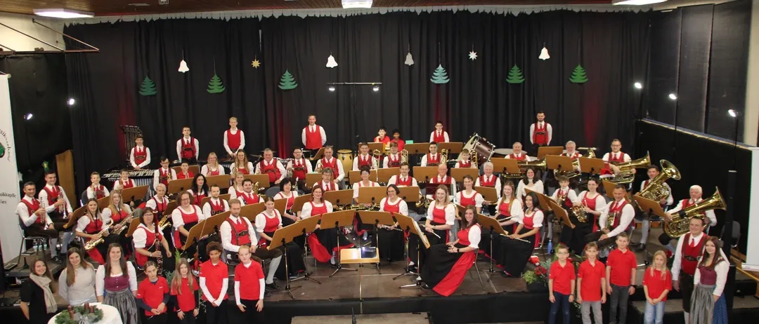 Ein großes Orchester mit verschiedenen Instrumenten tritt auf einer Bühne auf. Die Bandmitglieder sind in roten und weißen Kostümen gekleidet. Die Bühne ist mit Weihnachtsbäumen und Lichtern geschmückt.