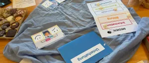 Ein Holztisch mit einem blauen Tuch, auf dem verschiedene Karten und Etiketten zu sehen sind, darunter lächelnde Gesichter, ein Dokument und eine blaue Mappe mit dem Wort 'Mittagessen'.