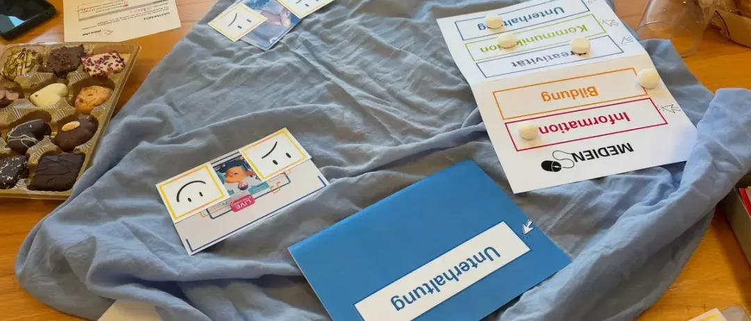 Ein Holztisch mit einem blauen Tuch, auf dem verschiedene Karten und Etiketten zu sehen sind, darunter lächelnde Gesichter, ein Dokument und eine blaue Mappe mit dem Wort 'Mittagessen'.