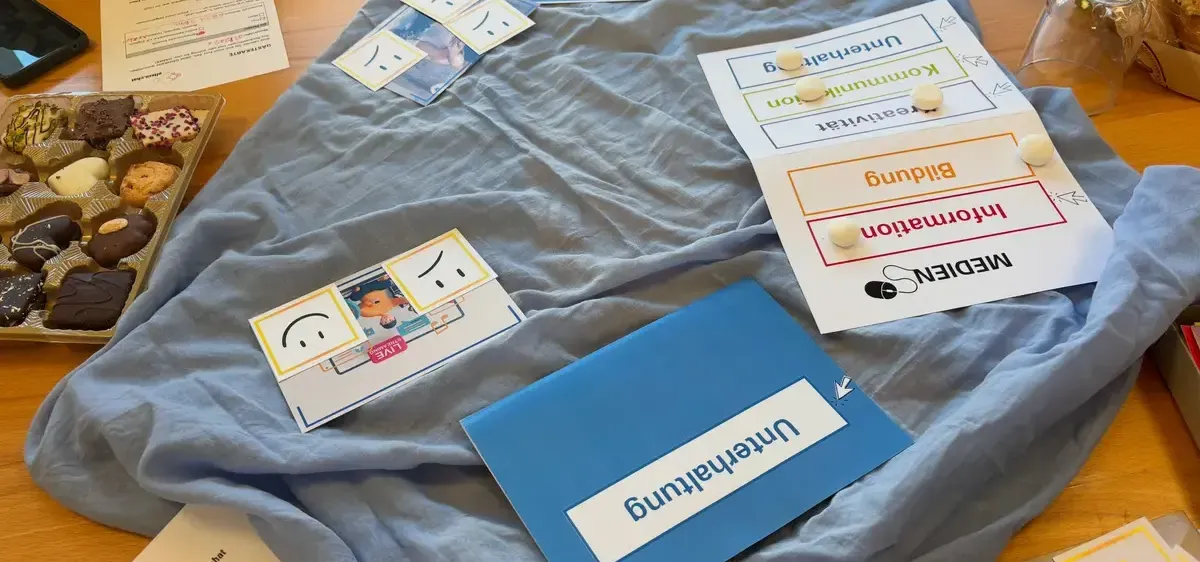 Ein Holztisch mit einem blauen Tuch, auf dem verschiedene Karten und Etiketten zu sehen sind, darunter lächelnde Gesichter, ein Dokument und eine blaue Mappe mit dem Wort 'Mittagessen'.