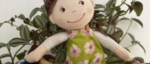 Eine handgefertigte Puppe mit lockigem braunem Haar und einem grünen Blumenkleid sitzt auf einer Holzoberfläche. Sie trägt rosa Schuhe und sitzt neben einem Paar blauer Musikglocken.