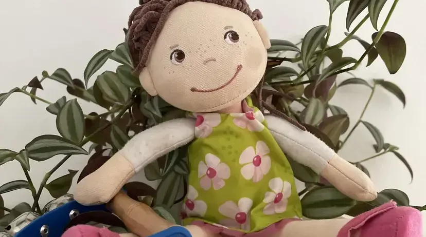 Eine handgefertigte Puppe mit lockigem braunem Haar und einem grünen Blumenkleid sitzt auf einer Holzoberfläche. Sie trägt rosa Schuhe und sitzt neben einem Paar blauer Musikglocken.