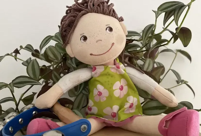 Eine handgefertigte Puppe mit lockigem braunem Haar und einem grünen Blumenkleid sitzt auf einer Holzoberfläche. Sie trägt rosa Schuhe und sitzt neben einem Paar blauer Musikglocken.