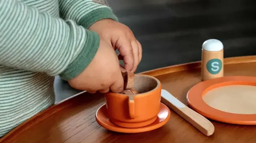 Ein Kind platziert Stücke von Lebensmitteln in einen orangefarbenen Becher, der auf einer Untertasse auf einem Holztisch steht.