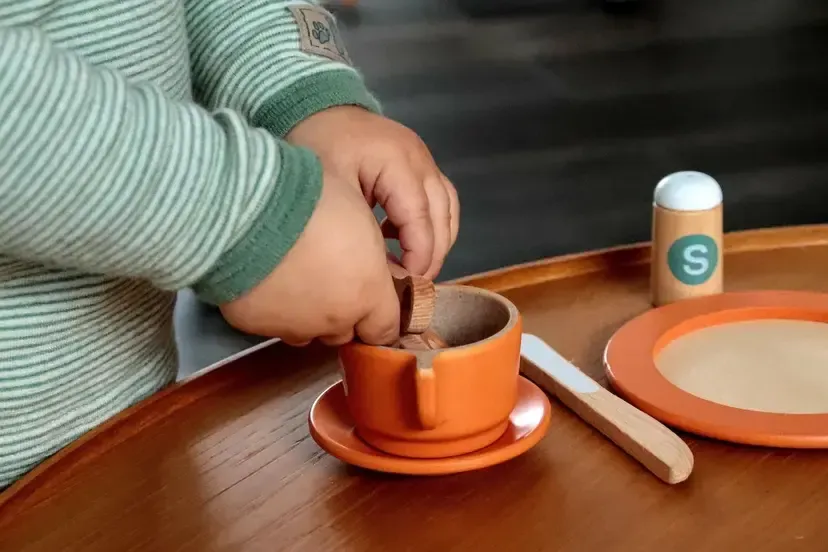Ein Kind platziert Stücke von Lebensmitteln in einen orangefarbenen Becher, der auf einer Untertasse auf einem Holztisch steht.