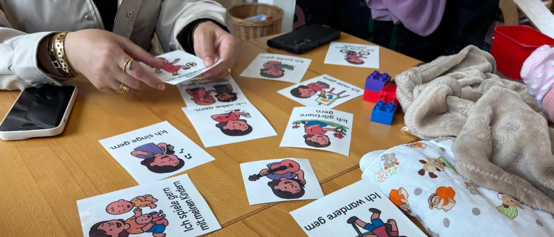Eine Person hält eine Karte mit einer Cartoon-Figur und Text. Mehrere Karten mit ähnlichen Illustrationen und Texten liegen auf einem Holztisch.