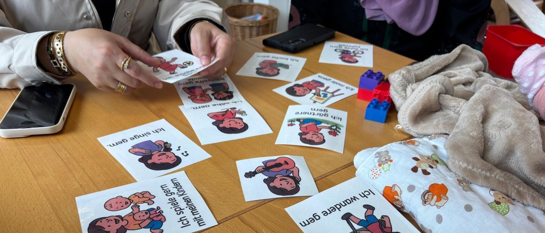 Eine Person hält eine Karte mit einer Cartoon-Figur und Text. Mehrere Karten mit ähnlichen Illustrationen und Texten liegen auf einem Holztisch.