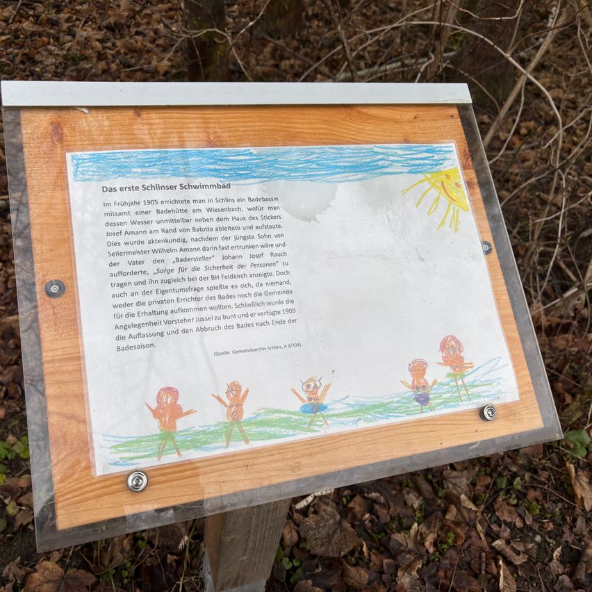 Ein Schild im Wald zeigt eine Zeichnung von Kindern, die schwimmen, und einen historischen Text auf Deutsch über das erste Schwimmbad in Schlins.