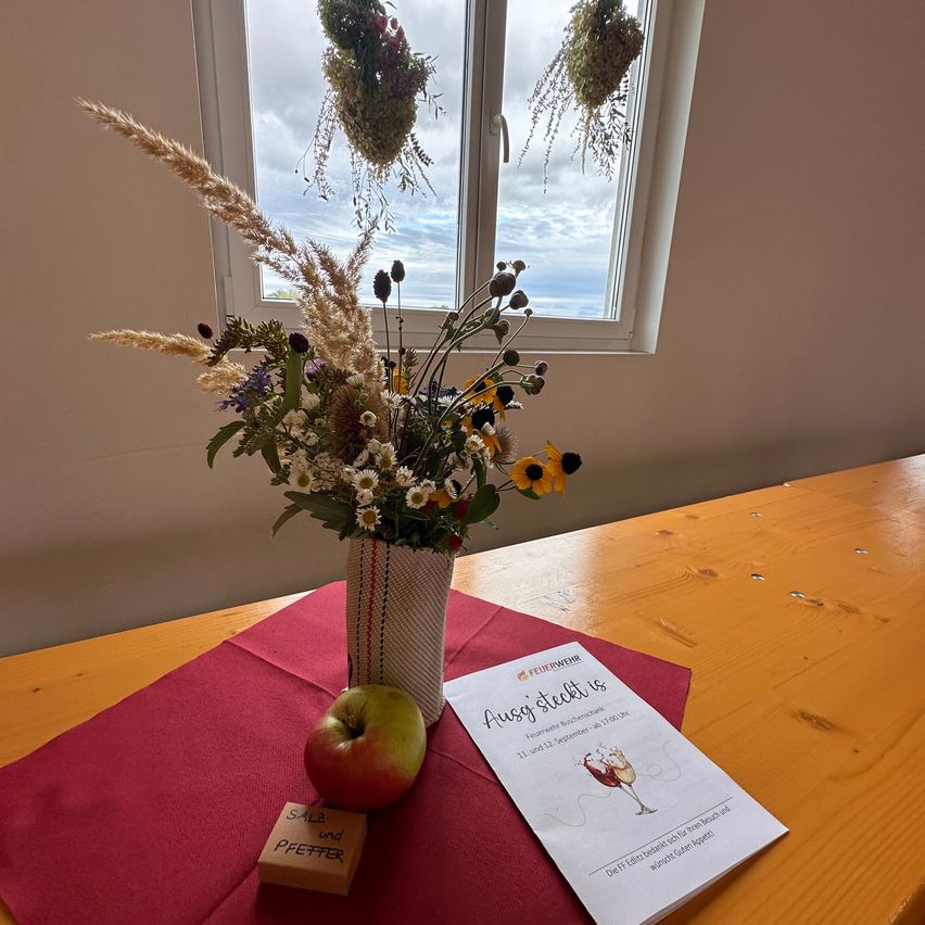 Ein Holztisch mit einem roten Tuch, einer Vase mit Wildblumen, einem roten Apfel und einem kleinen Holzkasten mit der Aufschrift 'Salz und Pfeffer'. Eine Einladungskarte mit dem Text 'Aussy steckli is' liegt auf dem Tisch. Das Fenster hinter dem Tisch bietet einen Blick auf den Himmel und Wolken.
