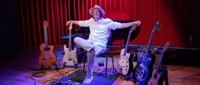 Ein Mann in einem Hut sitzt auf einem Stuhl auf der Bühne und posiert für ein Foto mit verschiedenen Gitarren vor ihm.