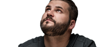 Ein Mann mit Bart trägt ein graues T-Shirt. Er schaut nach oben und posiert für ein Foto vor schwarzem Hintergrund.