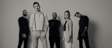 Ein Schwarz-Weiß-Bild zeigt fünf Menschen in einer Reihe, wahrscheinlich eine Band, die gegen eine schlichte weiße Wand stehen. Der Mann in der Mitte trägt Hosenträger und einen Schnurrbart.