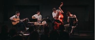 Vier Musiker spielen auf der Bühne mit Cello, Gitarre und Akkordeon. Ein Schlagzeuger sitzt vor einem Schlagzeug, mit Scheinwerfern, die die Szene beleuchten. Das Publikum beobachtet aus den Schatten.