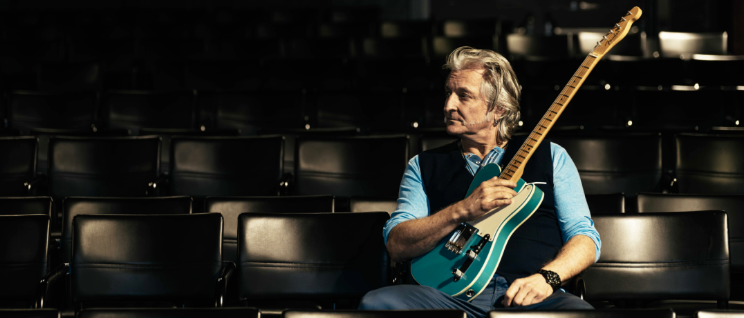 Ein älterer Mann sitzt in einem schwach beleuchteten Theater mit einer türkisfarbenen Gitarre und schaut zur Seite, möglicherweise in Gedanken versunken.