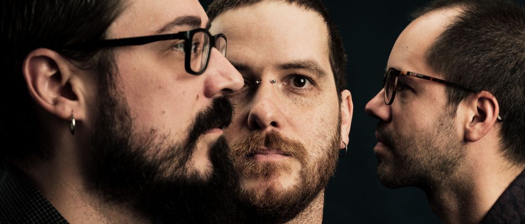 Drei Männer mit Bärten und Brillen, jeder starrt nach vorne. Der linke Mann hat einen Piercing und der mittlere Mann hat ein Muttermal auf der Nase.