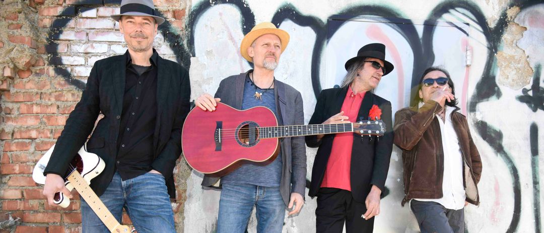 Vier Männer stehen vor einer mit Graffiti bedeckten Wand, zwei mit Gitarren und zwei mit einer Zigarette. Der Mann in der Mitte hält eine Gitarre, während der Mann auf der rechten Seite raucht.