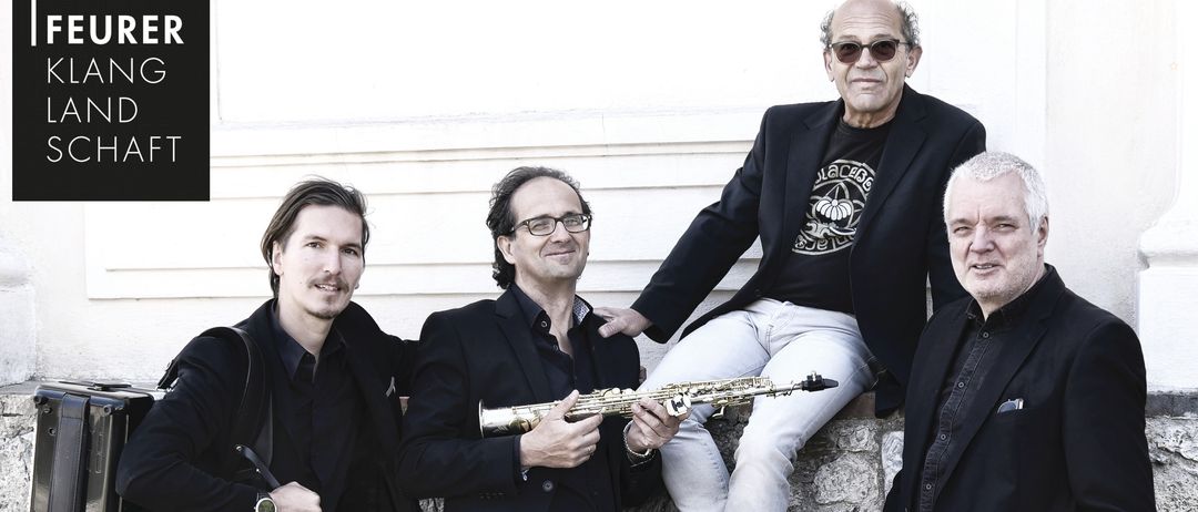 Vier Männer in schwarzen Anzügen posieren für ein Foto. Einer sitzt auf einer Steinmauer, ein anderer hält ein Saxophon und die beiden anderen halten eine Gong und eine Aktentasche.