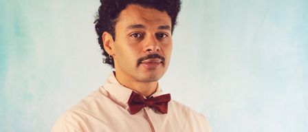 Ein Mann mit Schnurrbart, der ein rosa Hemd und eine rote Fliege trägt, posiert für ein Foto vor blauem Hintergrund.