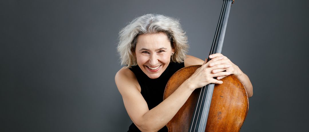Eine Frau mit weißen Haaren sitzt und lächelt, während sie ein Cello hält. Sie trägt ein schwarzes ärmelloses Oberteil. Das Cello hat einen braunen Korpus, einen schwarzen Hals und einige schwarze Wirbel.