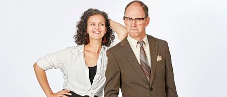 Eine Frau mit lockigem Haar lächelt und posiert mit der Hand am Kopf. Neben ihr steht ein älterer Mann in einem Anzug, der eine Brille und eine Krawatte trägt.